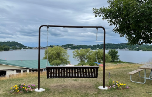 A swing set overlooking a calm lake, surrounded by trees and flowers under a cloudy sky.