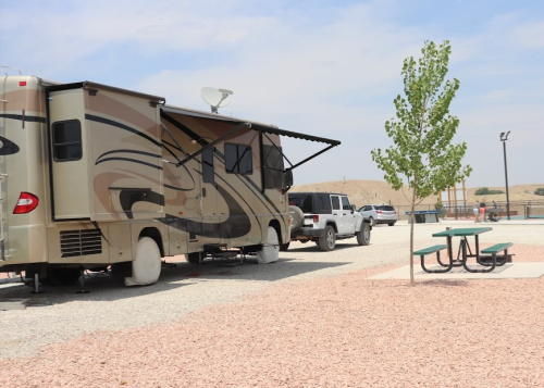 An RV parked beside a picnic table and tree in a gravel area, with vehicles in the background and a clear sky.