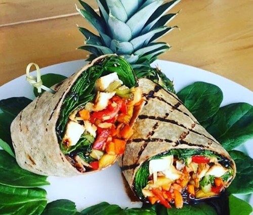 A colorful wrap filled with vegetables and chicken, served on a bed of greens with a pineapple in the background.