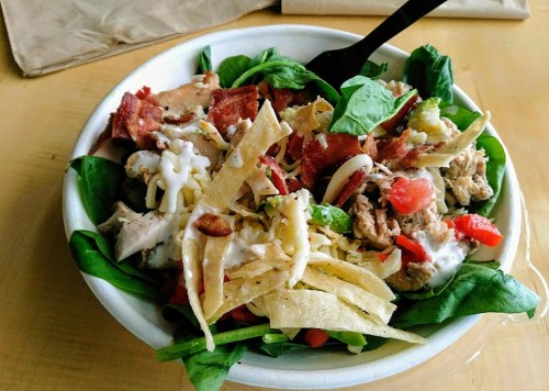 A colorful salad bowl with spinach, grilled chicken, bacon, cheese, tomatoes, and tortilla strips, topped with dressing.