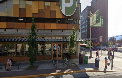 A modern building with a large green sign, outdoor seating, bicycles parked, and pedestrians on a sunny street.