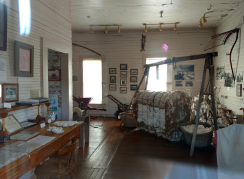 Interior of a historical museum room featuring cotton bales, vintage tools, and framed photographs on the walls.