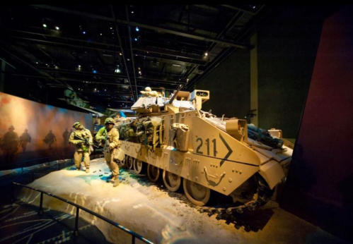 A military vehicle displayed in a museum, with soldiers in uniform nearby, set in a dimly lit environment.