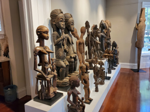 A display of various wooden sculptures and figures arranged along a white shelf in a gallery setting.