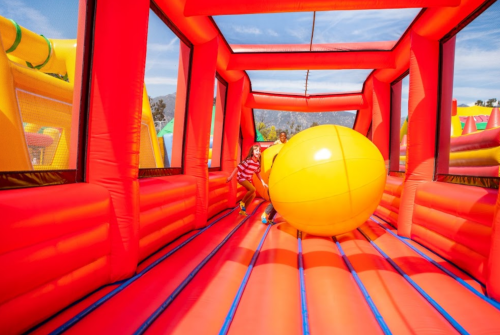 Two people play with a large yellow ball inside a bright red inflatable structure on a sunny day.