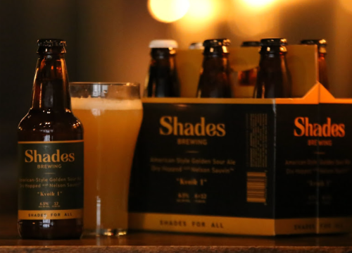 A bottle and glass of Shades Brewing's "Kwell 1" sour ale, with a six-pack in the background.