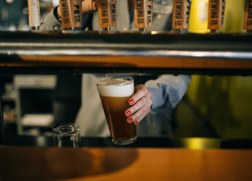 A hand with red nails pours a beer into a glass at a bar, with taps visible in the background.