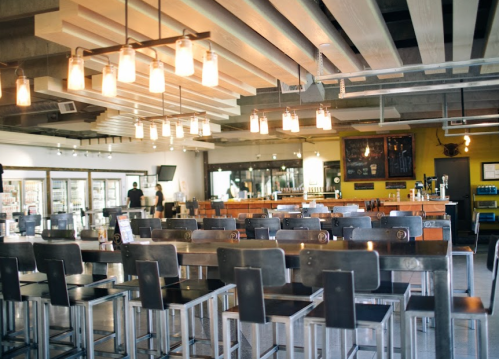 Modern restaurant interior with metal bar stools, wooden accents, and pendant lighting, featuring a spacious layout.