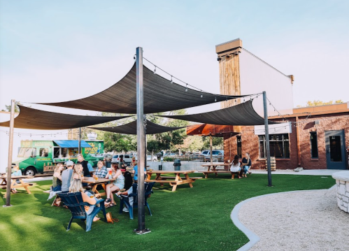 Outdoor dining area with picnic tables, shade sails, and a food truck in a lively atmosphere.