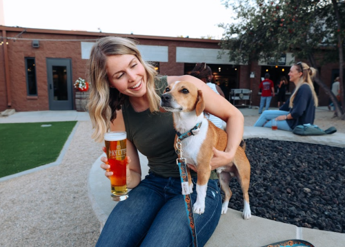 A woman smiles at her dog while holding a beer in a lively outdoor setting.