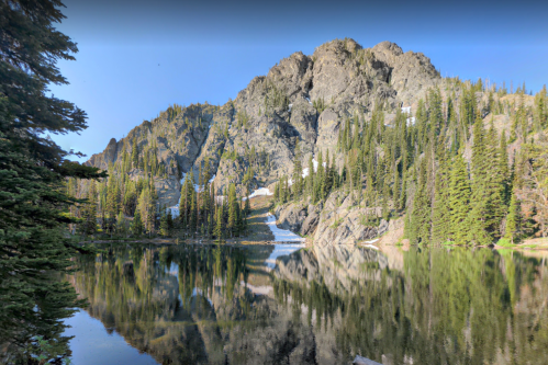 A serene mountain landscape with a clear lake reflecting rocky cliffs and lush green trees under a blue sky.