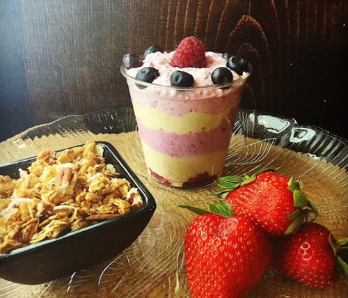A layered fruit parfait with yogurt, topped with berries, alongside a bowl of granola and fresh strawberries on a plate.