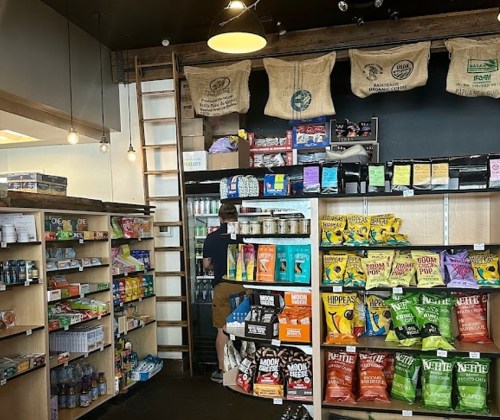 A cozy store interior with shelves stocked with snacks, drinks, and goods, featuring a ladder and hanging burlap sacks.