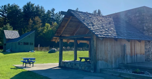 A wooden pavilion with a stone base and picnic table, surrounded by green grass and trees in the background.