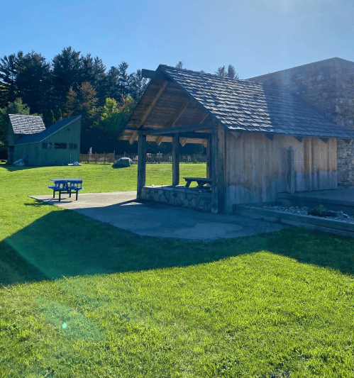 A sunny outdoor area with a wooden shelter, stone wall, and green grass, surrounded by trees and picnic tables.