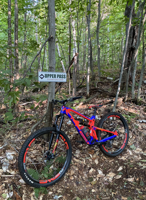 A brightly colored mountain bike leans against a tree in a forest, next to a sign that reads "Upper Pass."