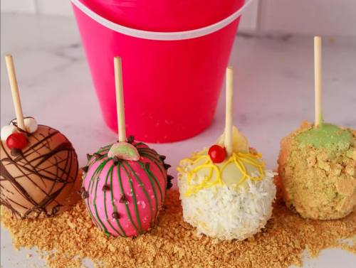 Four decorated caramel apples on a marble surface, with a pink bucket in the background and crushed graham crackers scattered.