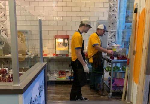 Two workers in yellow shirts and hats prepare food in a small kitchen with a popcorn machine and colorful ingredients.