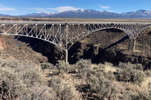A large metal bridge spans a deep canyon, with snow-capped mountains in the background and sparse vegetation below.