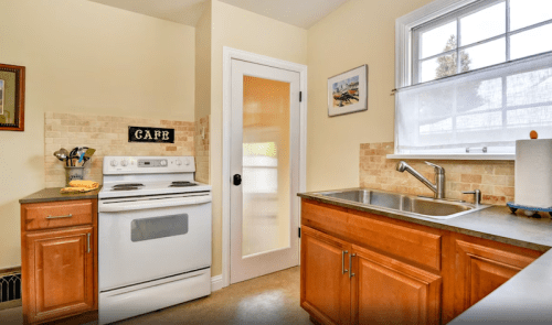 A bright kitchen featuring a white stove, wooden cabinets, a sink, and a door leading to another room.