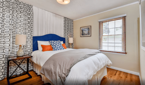 Cozy bedroom with a blue headboard, orange and white pillows, patterned curtains, and wooden floors.