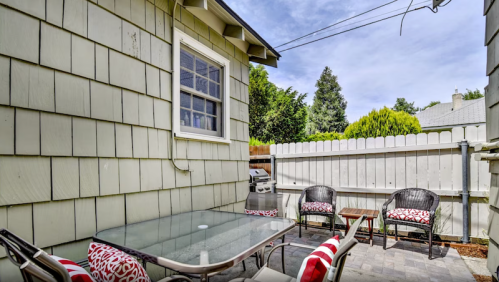 A cozy outdoor patio with a glass table and chairs, surrounded by greenery and a wooden fence.
