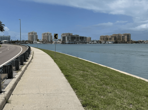 A scenic view of a waterfront path with buildings in the background under a clear blue sky.