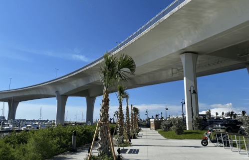 A modern bridge arches over a marina, with palm trees lining a walkway under a clear blue sky.
