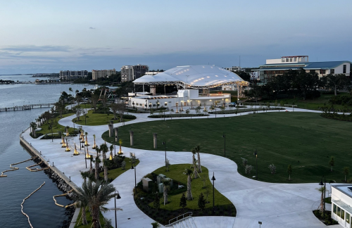 A waterfront park with a large pavilion, palm trees, and a winding path along the water's edge at dusk.