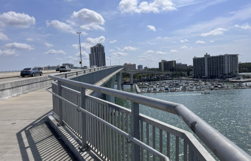 A bridge with a railing over a waterway, leading to buildings and boats in the distance under a partly cloudy sky.