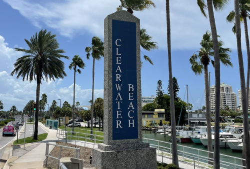 Sign for Clearwater Beach surrounded by palm trees and a marina under a blue sky.