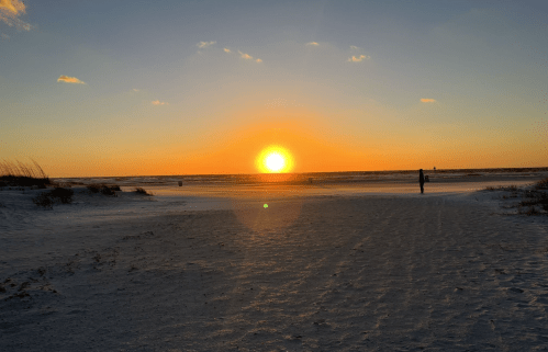 A serene beach at sunset, with a vibrant orange sun setting over the horizon and a silhouette of a person in the distance.
