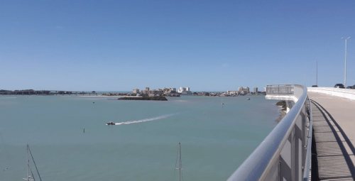 A view from a bridge overlooking a calm turquoise bay with a small boat and distant buildings on the shore.