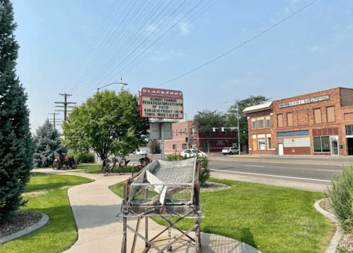 A street view featuring a sign for Blackfoot, Idaho, with nearby buildings and greenery along the sidewalk.