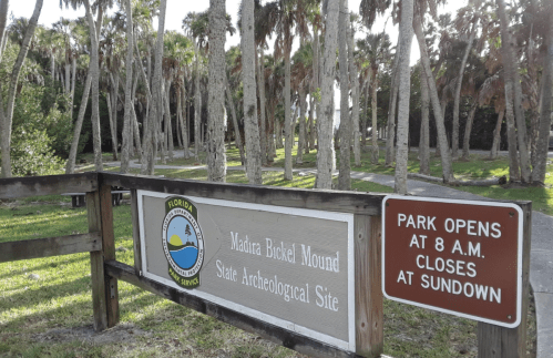 Sign for Madira Bickel Mound State Archeological Site, with palm trees in the background and park hours displayed.