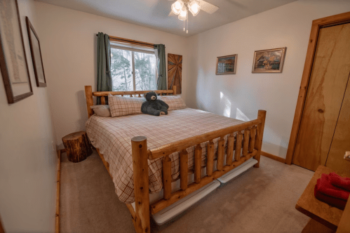 Cozy bedroom with a wooden bed, plush bear, and natural light from a window, featuring rustic decor and soft carpeting.