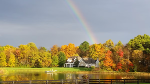 A serene landscape featuring a house by a lake, surrounded by vibrant autumn trees and a rainbow in the sky.