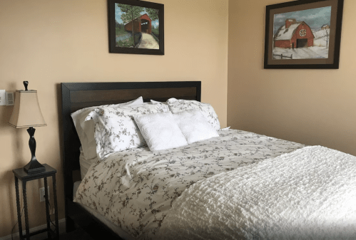 Cozy bedroom with a bed covered in floral bedding, a lamp, and two framed barn paintings on the wall.