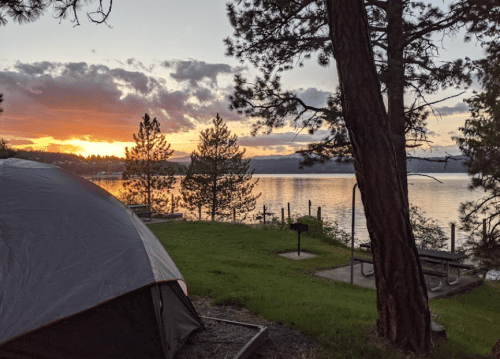 A serene lakeside view at sunset, featuring a tent, trees, and picnic areas by the water.