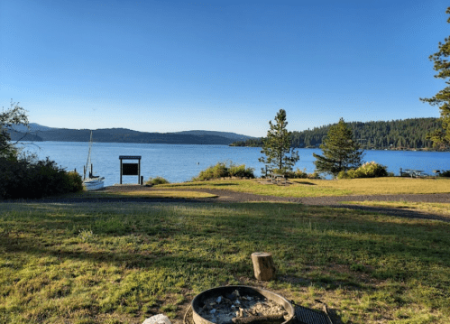 A serene lake view with trees, a fire pit in the foreground, and a clear blue sky.