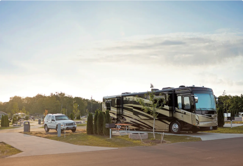 A luxury RV parked in a campsite with a white SUV nearby, surrounded by trees and a sunset sky.