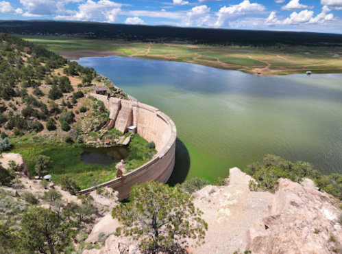 Aerial view of a curved dam surrounded by green water and lush landscape under a blue sky with scattered clouds.