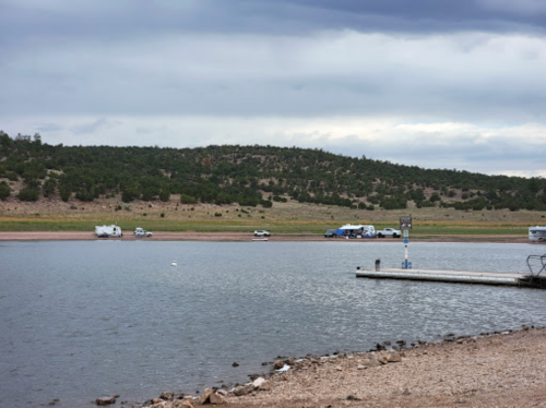 A serene lake scene with campers along the shore and a dock extending into the water, surrounded by hills and cloudy skies.