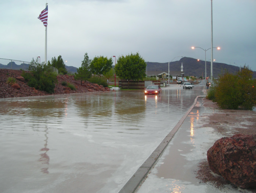 A flooded street with a car navigating through water, surrounded by trees and a flagpole in the background.