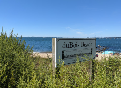 Sign for duBois Beach with a view of the water and sandy shore in the background, framed by greenery.