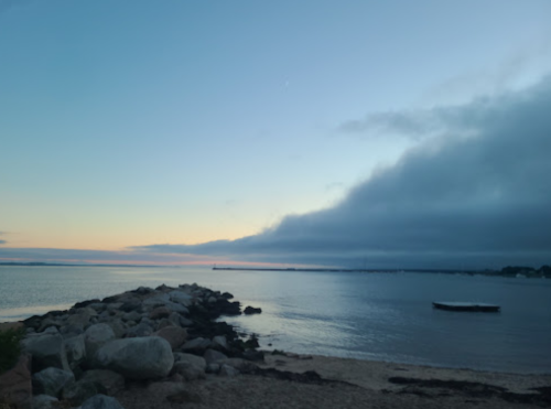 A serene beach scene at dusk, featuring calm waters, a rocky shoreline, and a cloudy sky with soft pastel colors.
