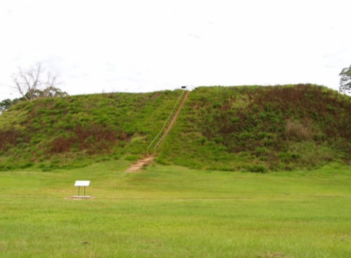 A grassy mound with a path leading up, surrounded by a green field and a sign at the base. Overcast sky above.