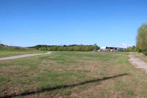 A wide, open field with a clear blue sky, bordered by trees and a building in the background.