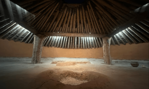 Interior of a traditional round structure with a wooden roof, featuring a central hearth and earthen floor.