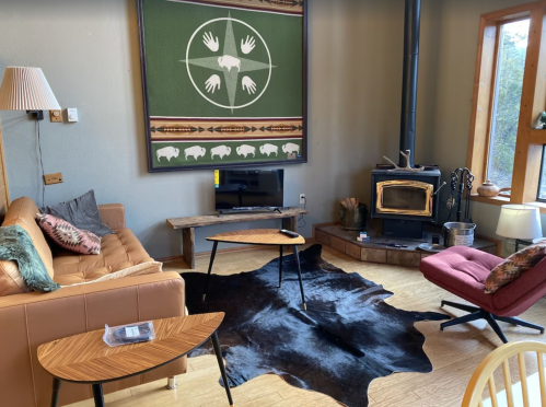 Cozy living room with a brown sofa, two coffee tables, a wood stove, and a large wall hanging featuring hands and buffalo.